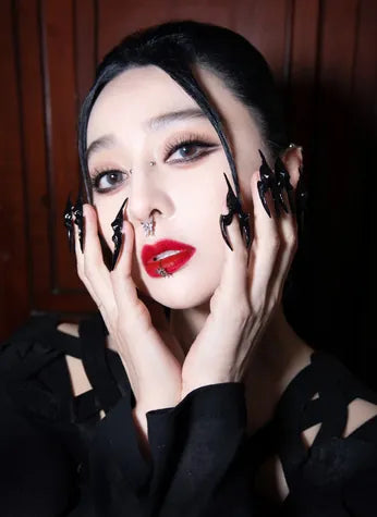 A portrait framed by hands in long black sculpted claw nails, a nose ring and red lips.
