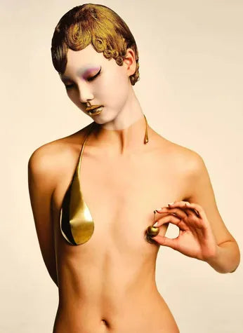 An editorial nude with a molten-gold breast piece and a gold sculpted cap.