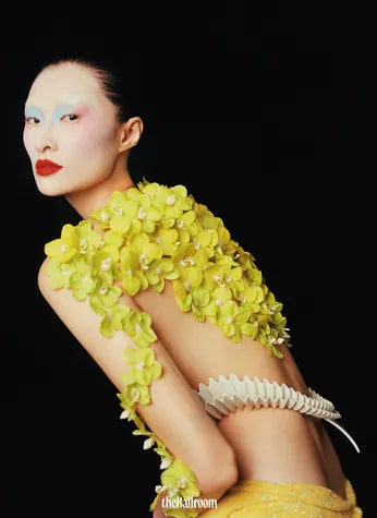 An editorial portrait in a coat of yellow orchids and a bone-white arm cuff.
