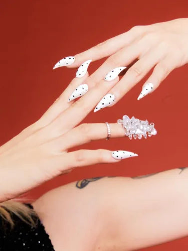 A hand against red in white polka-dot Dragon nails, a crystal cluster ring on one finger.