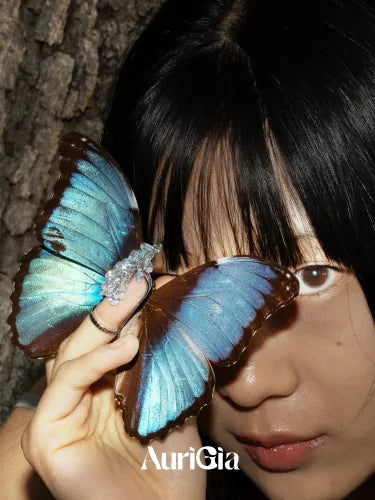 The model half hidden behind a blue morpho butterfly, a Glint ring on the finger holding it.